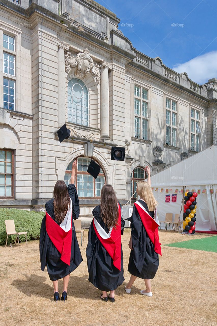 Back side view of three happy girls wearing dresses throwing academic caps in yard of Cardiff University