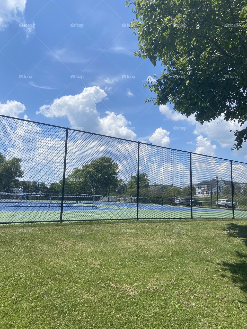 People playing tennis on blue tennis courts against the backdrop of white clouds, brilliant blue sky and green grass and trees. Colors and sport of summer!