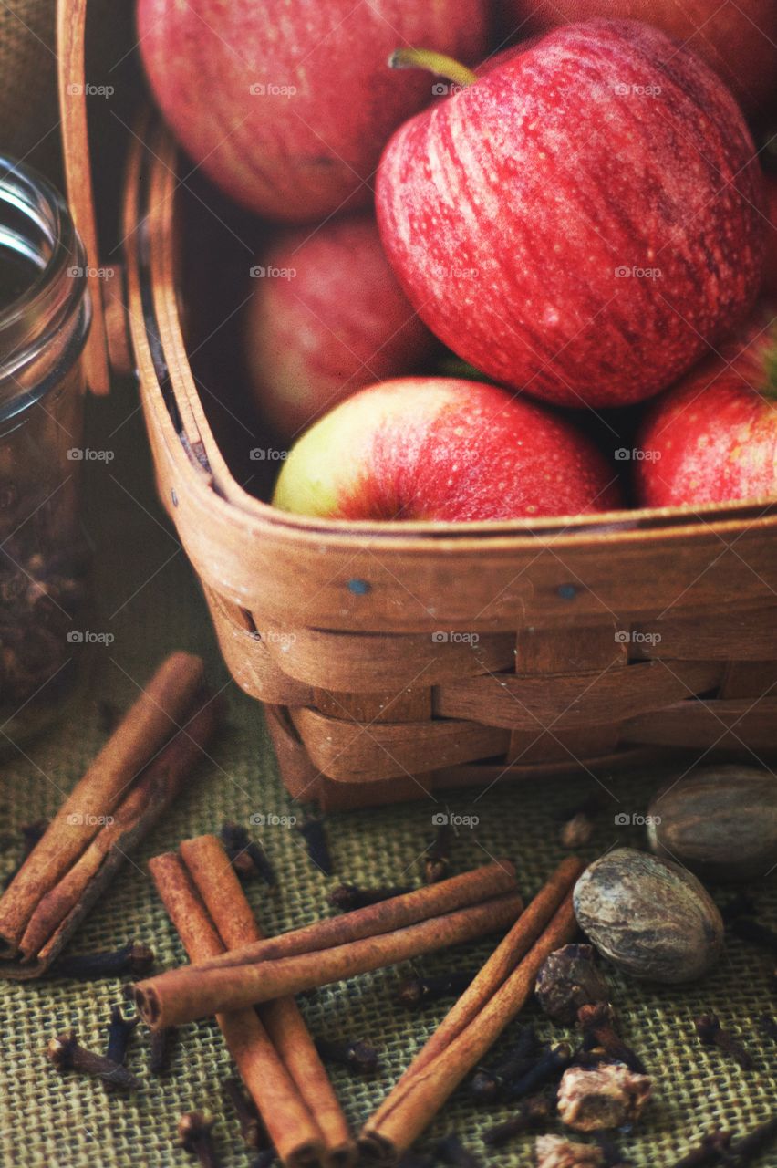 Cinnamon sticks, whole nutmeg, whole cloves, a small glass jar, and apples in a basket on burlap