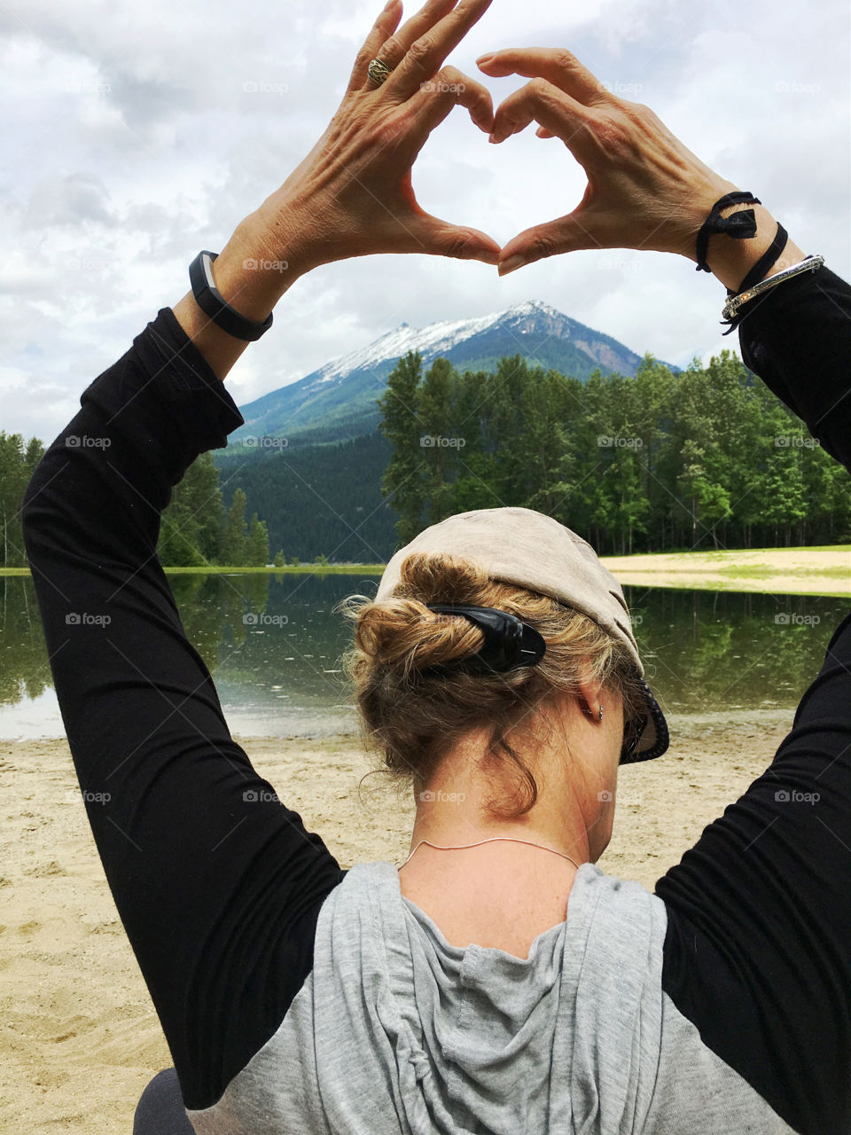 Blonde Woman in cap sitting in foreground of backdrop of Canada's snowy white Rocky Mountains and making heart sign with hands and arms
