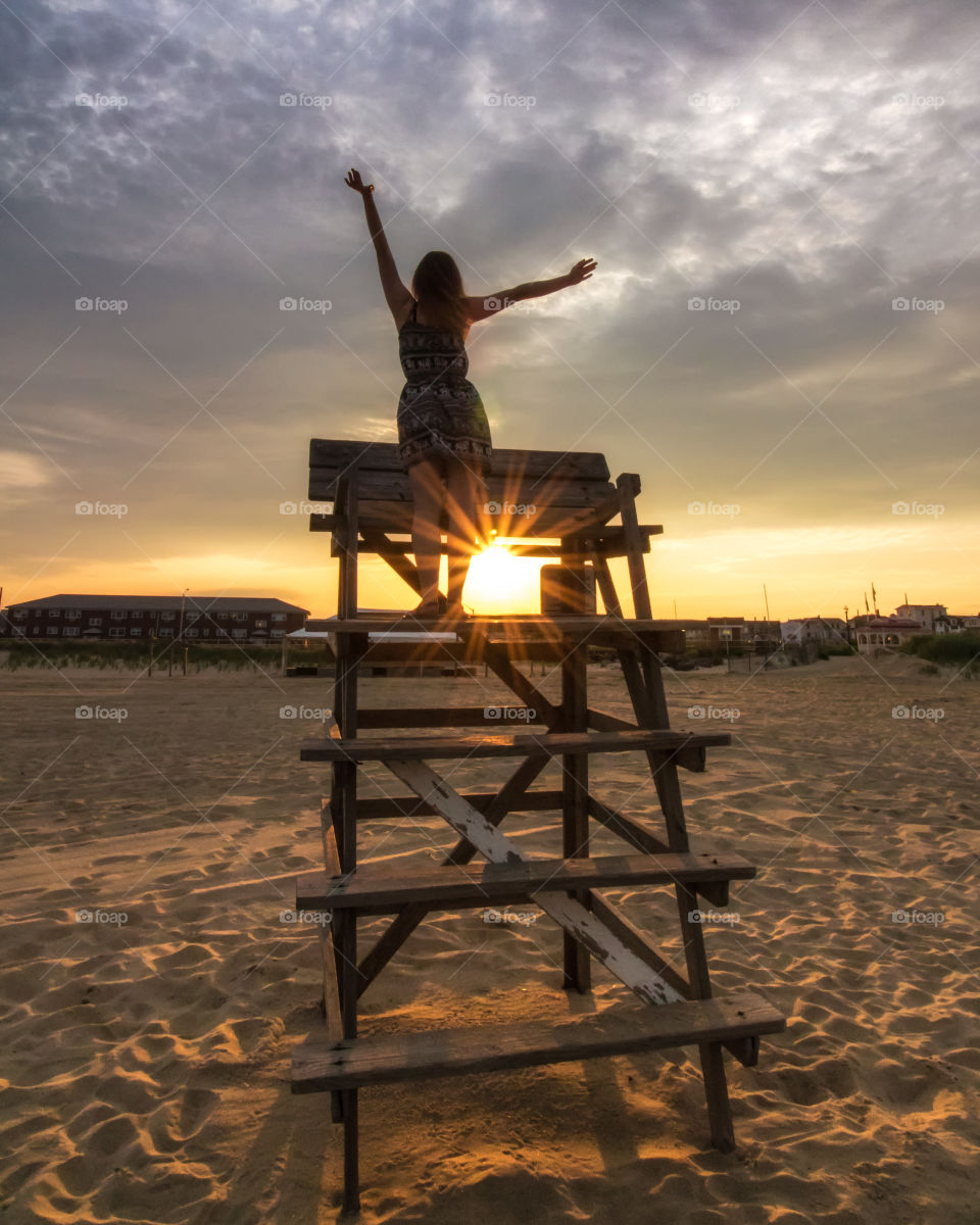 Girl standing on a lifeguard tower at the beach, taking in a beautiful vibrant sunset. Happy joyful pose .