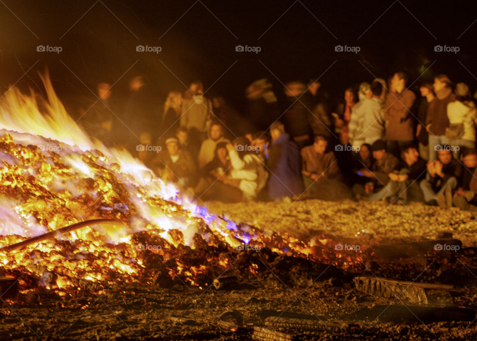 A large bonfire burns on the beach as a group of people sit around it