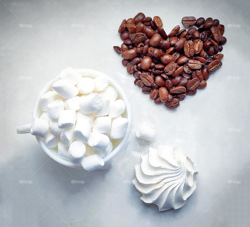 White colour. On a white background stands a white cup with a white marshmallow, a white marshmallow in the shape of a seashell and brown coffee beans in the shape of a heart. View from above