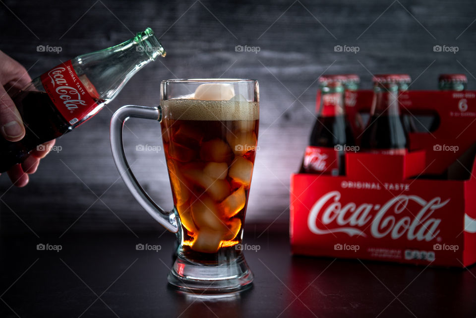 Person's hand pouring Coca-cola from a bottle into a tall backlit glass of Coca-cola