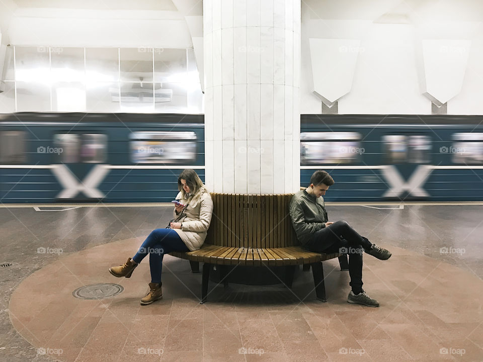 Young man and young woman using mobile phones in front of moving subway train 