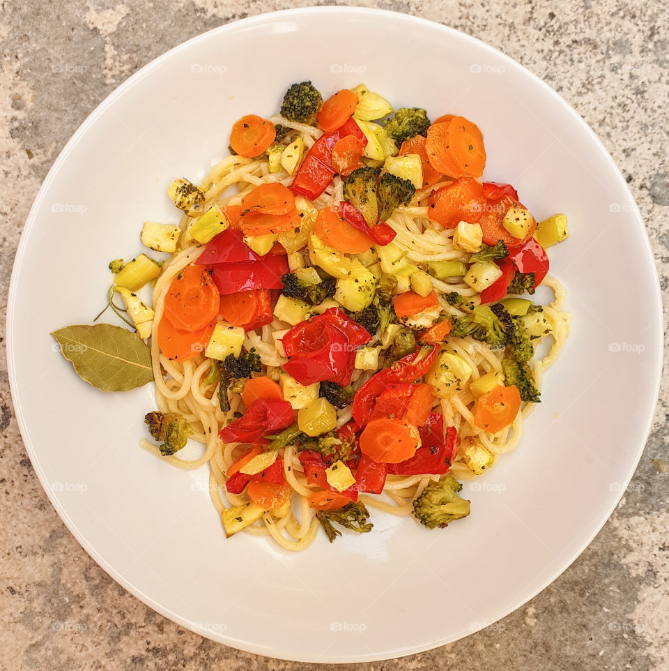 Spaghetti with baked seasonal colorful vegetables with bay leaf in a large white plate on a textured stone background. Healthy vegetarian food. Italian food