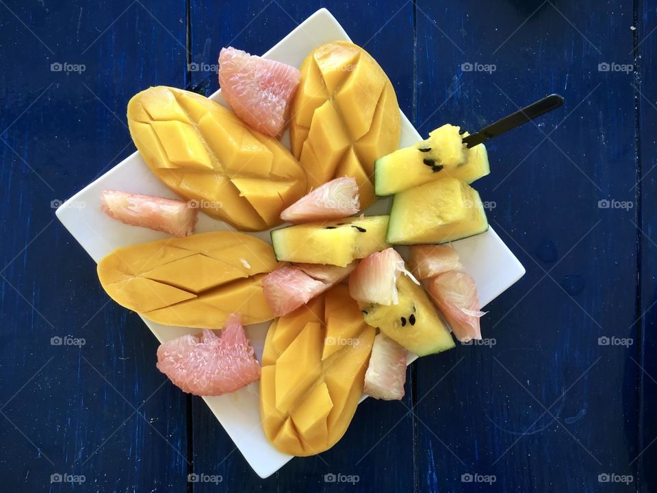 Variety of fruits on a white plate with blue table. Mango, watermelon, and grapefruit.