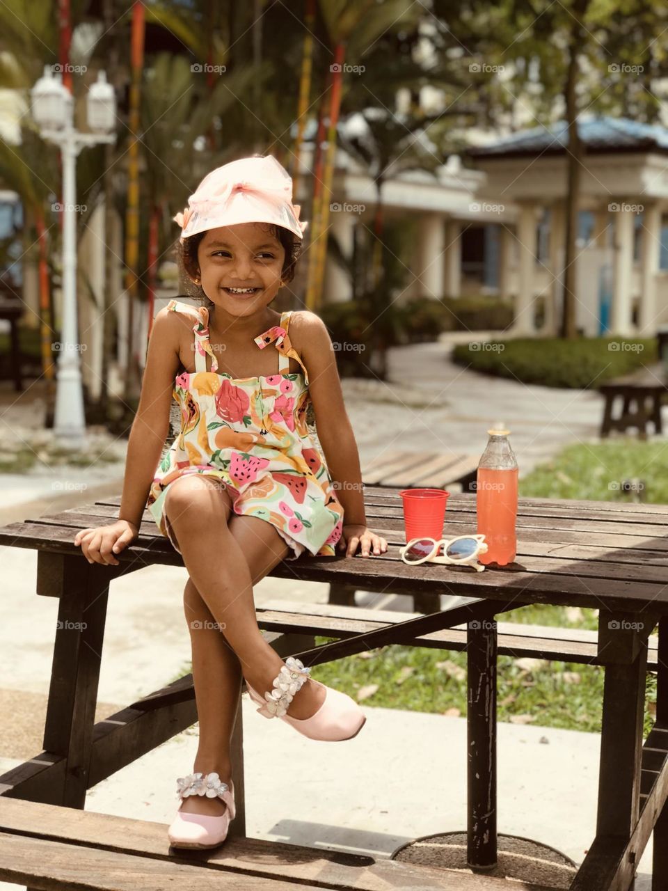 Baby girl on summer outfits sits over  outdoor table along with cool drink and cooling glasses and she has a adorable smile at her face 😍