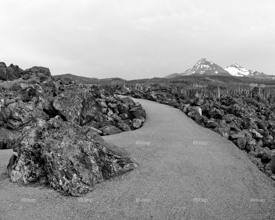 An asphalt pathway high up in Oregon's Cascade Mountains leads through rugged lava fields with two of the Three Sisters in the background. 