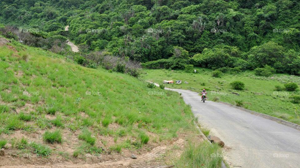 Lone rider - young girl riding motorbike on narrow winding road between green hills and bush in rural Transkei South Africa near Port St Johns