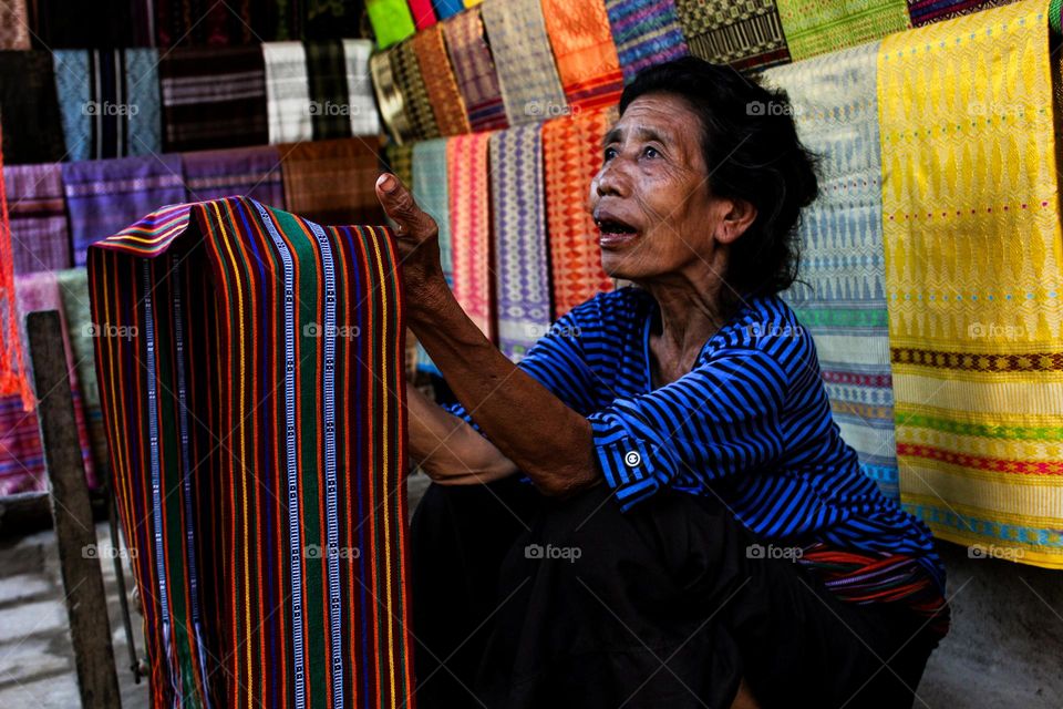 Portrait of an old woman sitting holding lombok woven cloth for sale as traditional souvenirs from Sade village, traditional cloth display in the background.