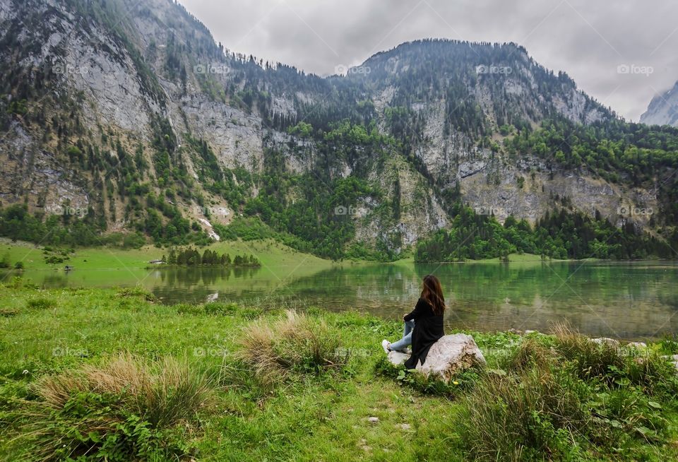 Back of people from behing, female person sitting on rock and enjoying peaceful scenic of lake and mountains in Germany in springtime