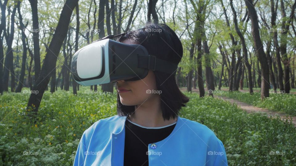 Portrait of trendy woman in blue coat using virtual reality glasses simulator in the park or forest at spring 