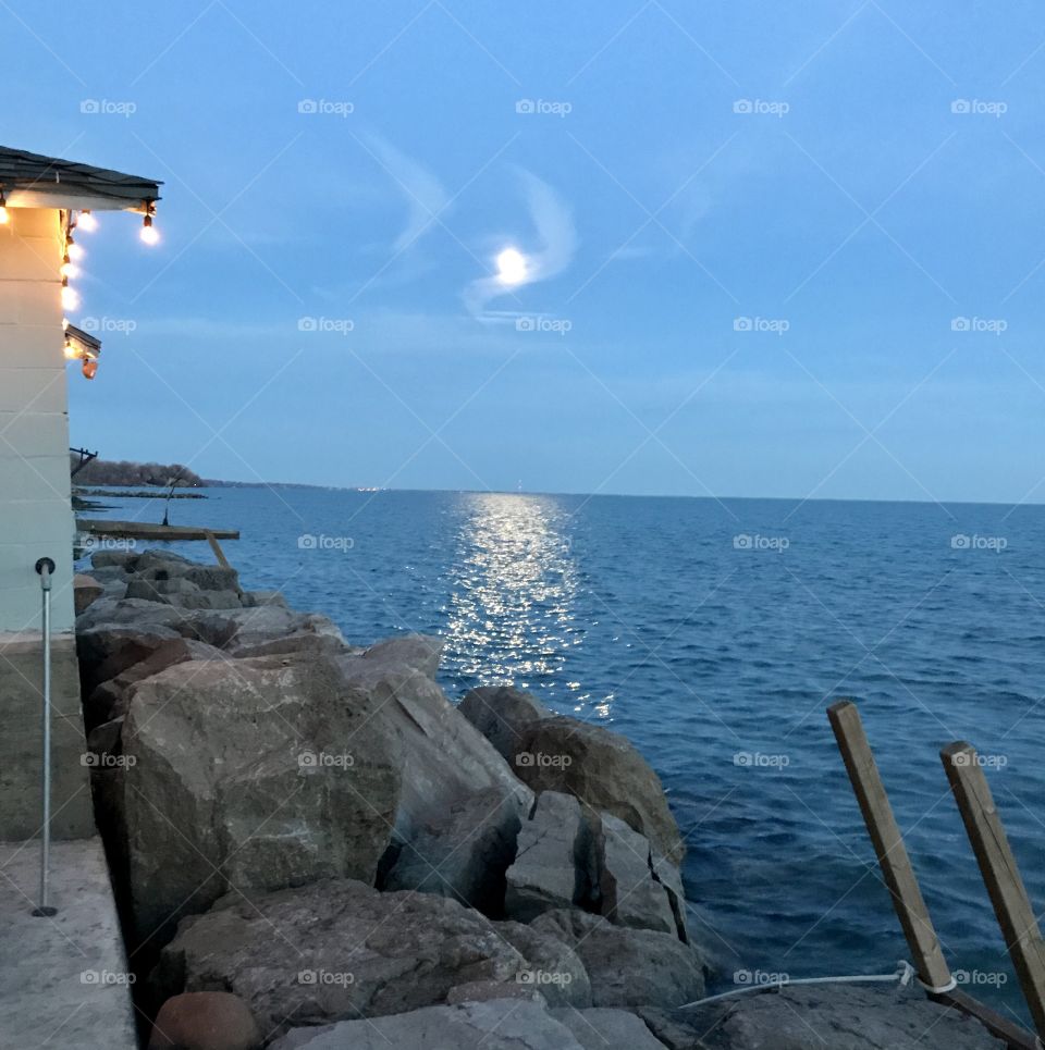 Tiny house with white twinkle lights on the edge of rock break wall on Lake Erie during moon rise.