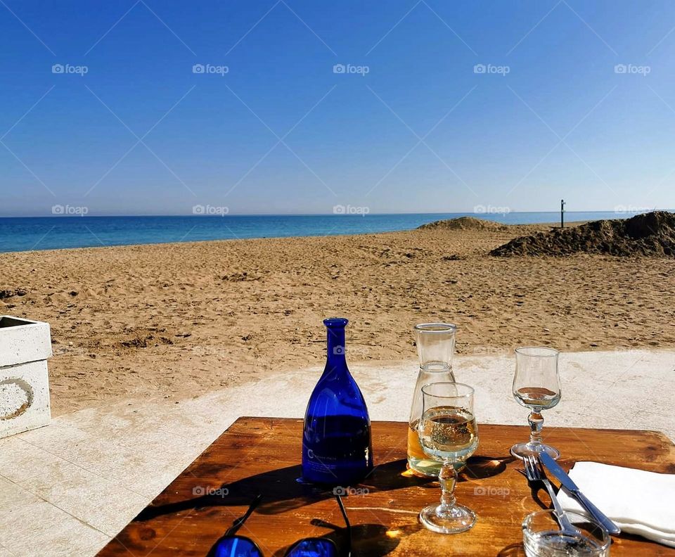 Sea.  Sandy shore.  The azure sea merges with the horizon.  In the foreground is a table with cutlery, two glasses of wine, bottles, sun glasses.  Romance