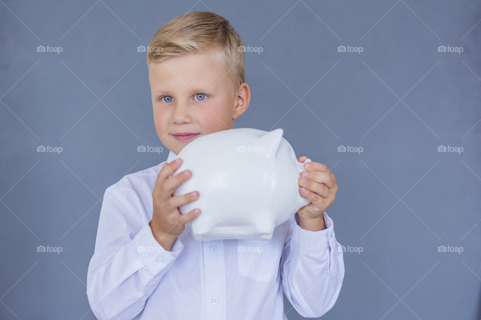 Beautiful smiling boy 6 years old in a white shirt with a bow tie stands in the studio on a blue background holds a piggy bank white pig  financial concept of children's pocket money