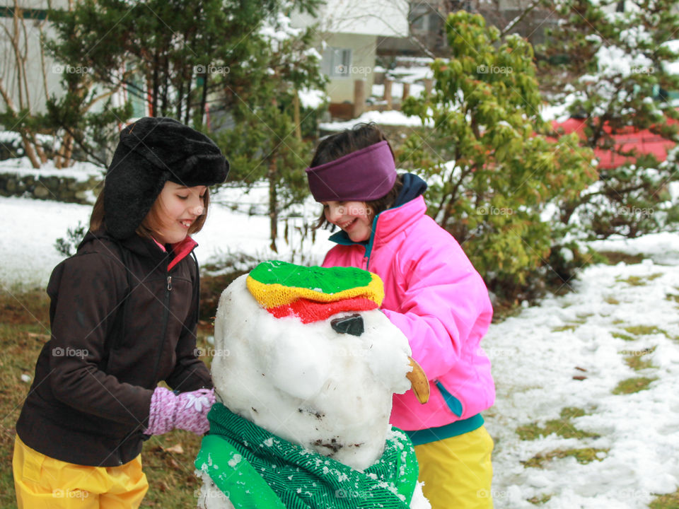 Two smiling sisters making a Reggae snowman complete with: a red, green & gold Rastafarian hat; a green scarf; a banana for a nose; and two smooth, black, beach stones for eyes. Yes, these are ‘Island’ girls! 🏝