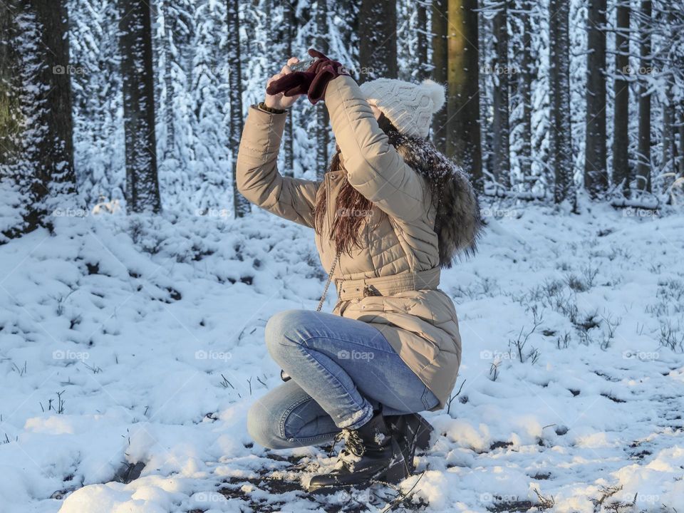 A beautiful young caucasian girl in a winter jacket and a hat with a pom-pom sitting on one knee shoots a snowy forest landscape on a mobile phone, side view close-up. Concept of winter landscape, girl blogger using technology.