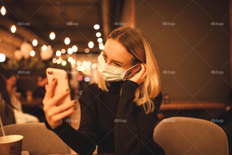 young woman sits in a mask.  blonde is sitting in a cafe and talking on the phone, quarantine time