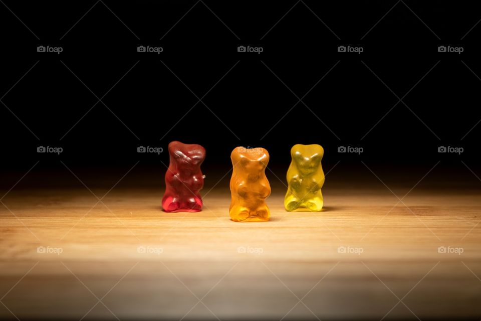 A portrait of three gummibears, the same but with other colors standing in the spotlight with darkness all around them.