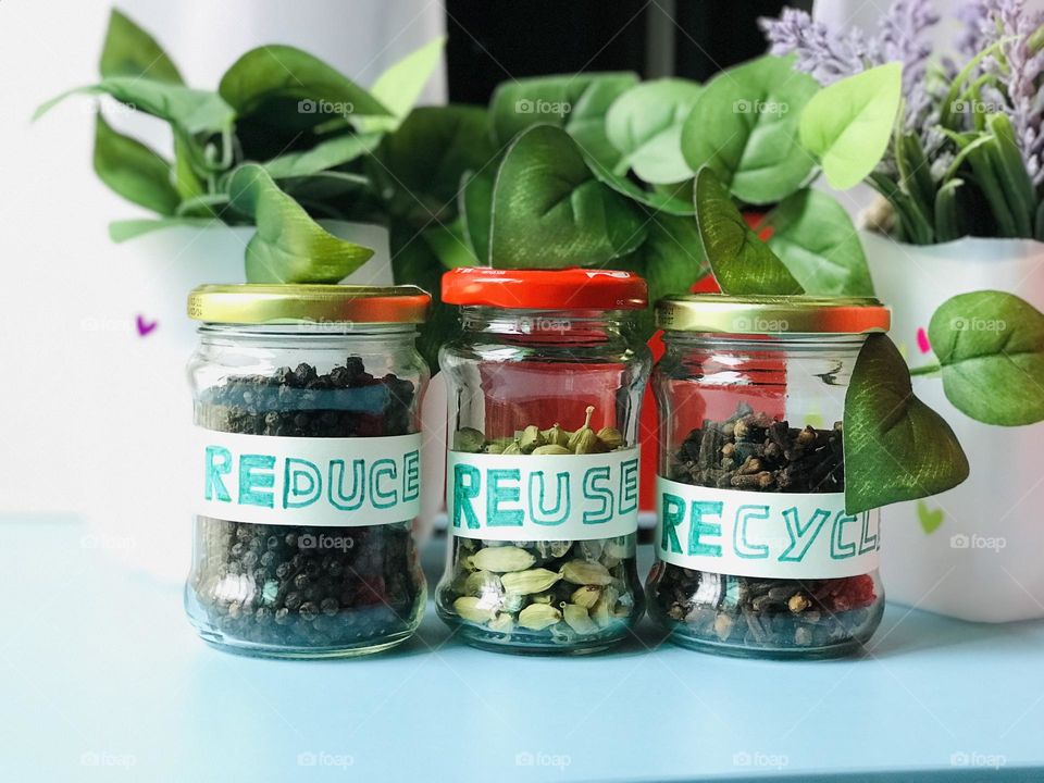 Three reusable glass containers with spices and labeled as Reduce reuse recycle and background covered with green plant leaves .