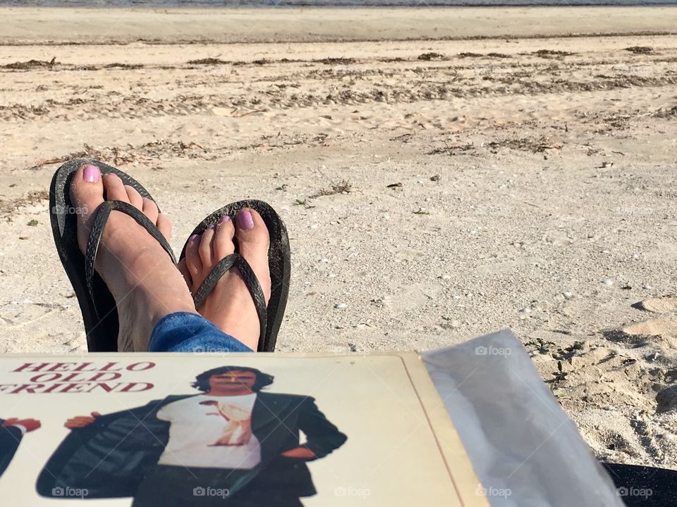 Woman (legs and feet only showing) laying on beach with vinyl record album on lap