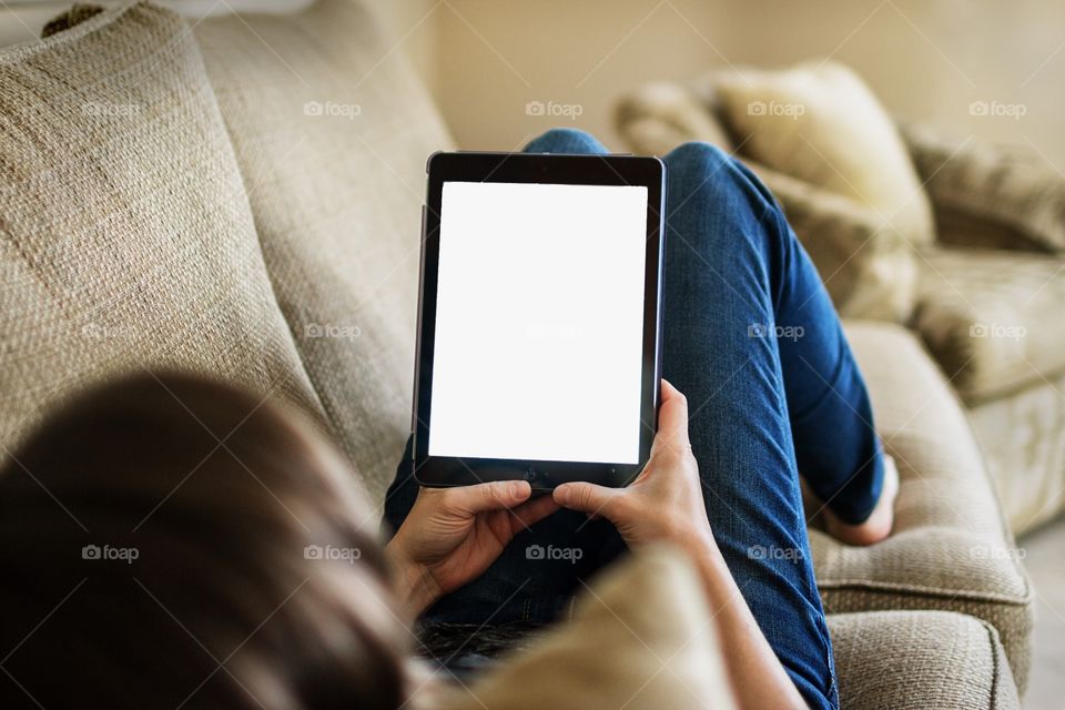 Woman holding iPad with blank white screen for mockup. Girl lounging on couch with iPad