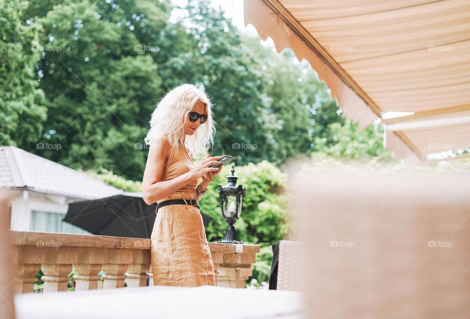 Adult elegant blonde woman in sunglasses and beige clothes using mobile phone in summer city street cafe