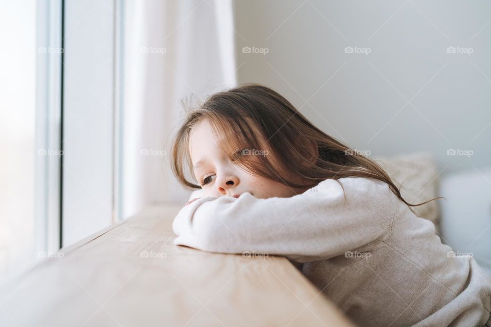 Cute bored thinking little girl with long hair in home clothes near window at the home
