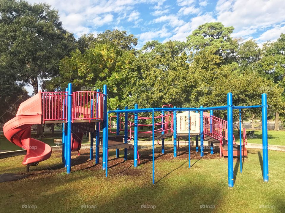 Play structure in a neighborhood park on a bright sunny day with blue sky and white clouds, with trees in the background 