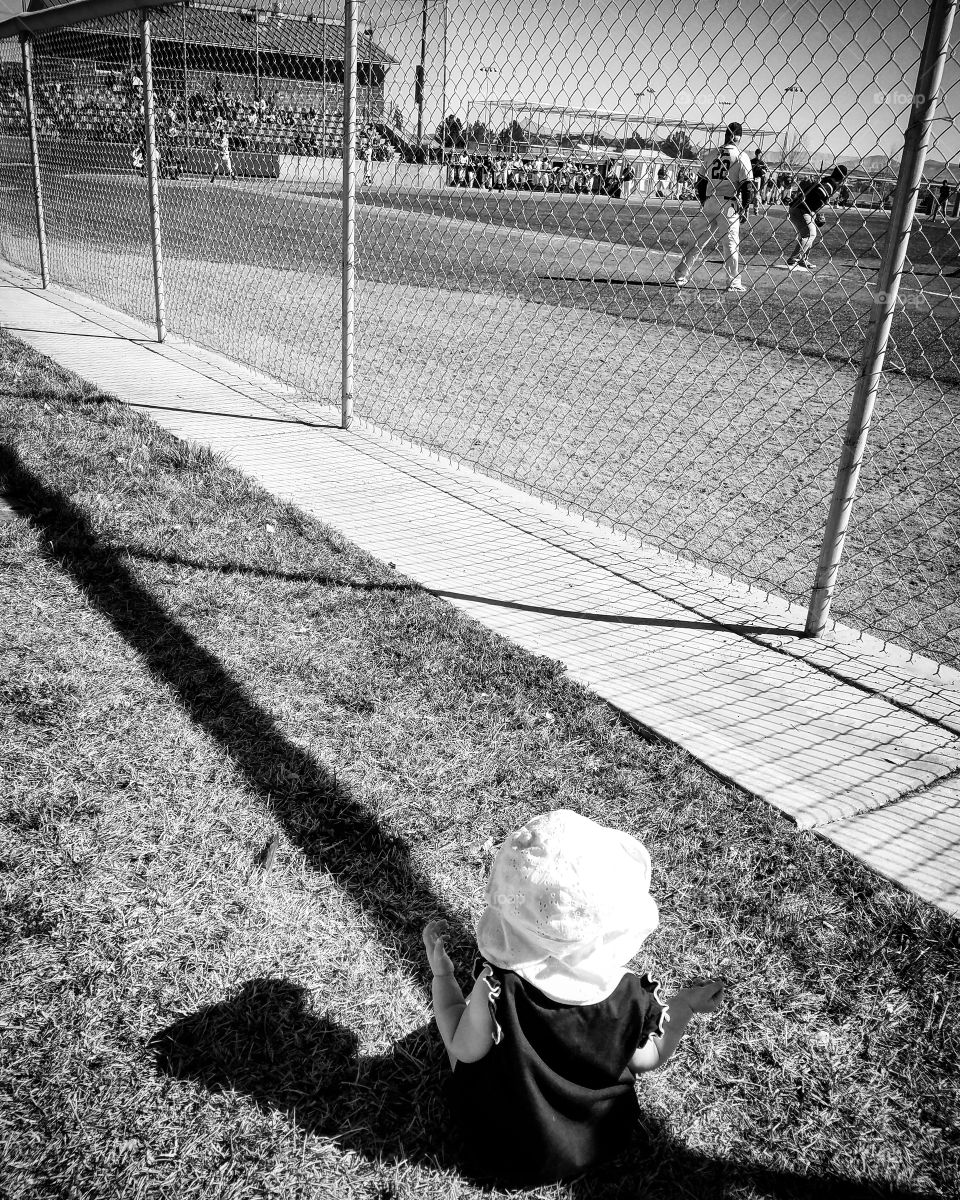 Baby girl sitting in grass in white sun hat watching high school baseball game through chain link fence