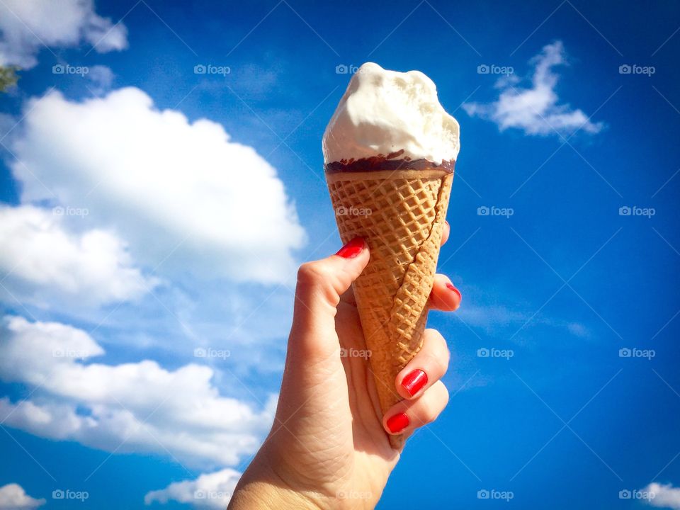 Woman's hand holding vanilla ice cream cone against blue sky with puffy white clouds