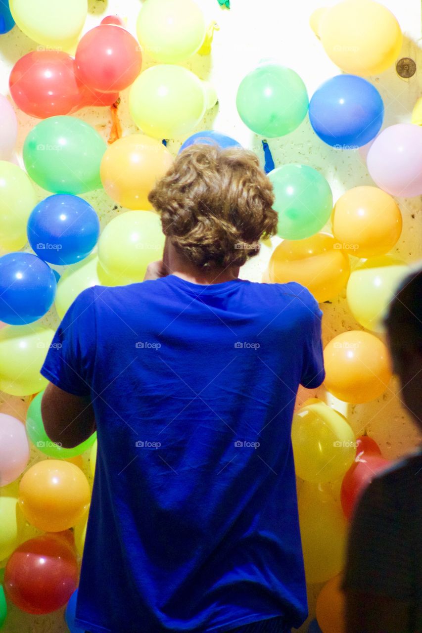 A young man in a royal blue t-shirt blows up brightly colored balloons in a carnival game booth