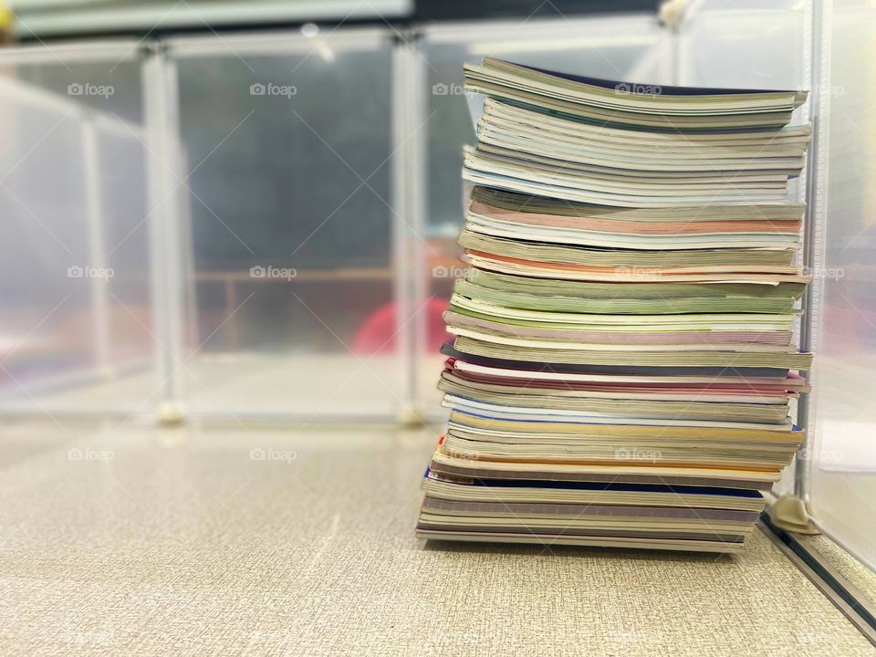 Several textbooks sit in an uneven pile on a clean and otherwise empty desk. Around the stack of books are plastic Covid protection walls. On the other side of the books is an empty chair. It is nighttime from the window outside.