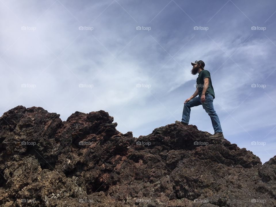 Bearded man in a baseball cap climbs a lava pile against a slate blue sky with wispy clouds