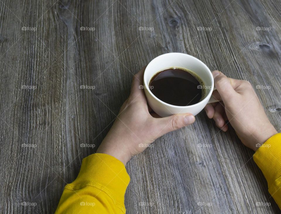 A man drinks coffee in the morning. Men's hands holding a white cup of coffee in a yellow jacket lie on a gray wooden surface, which is used as a background or a surface with incident light.