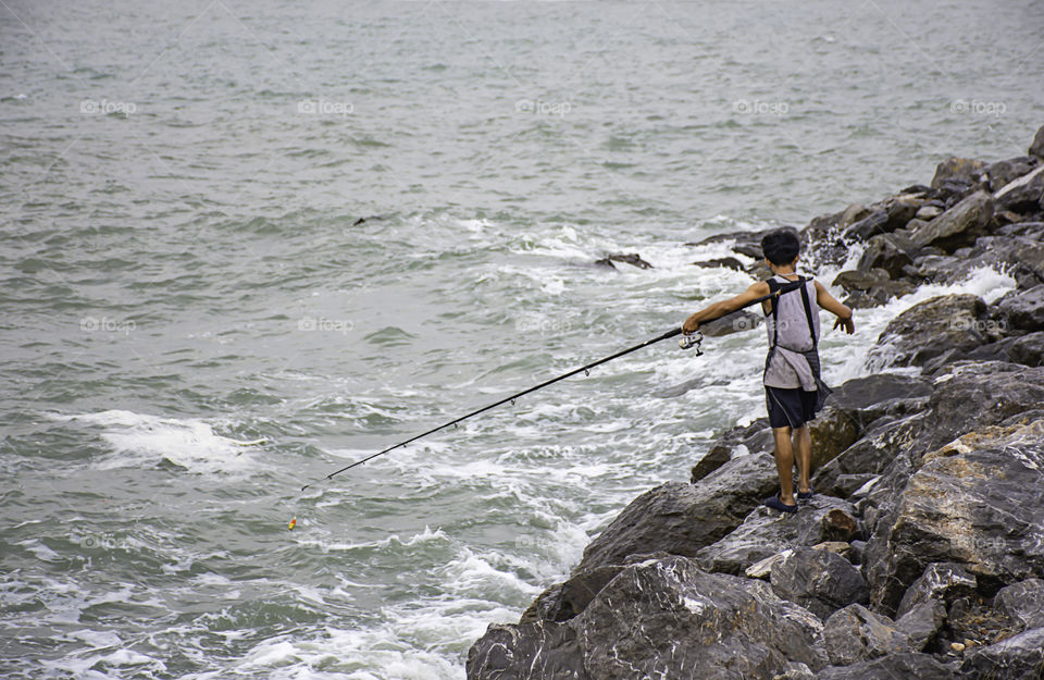 A man holding a fishing rod on the rock by the sea.