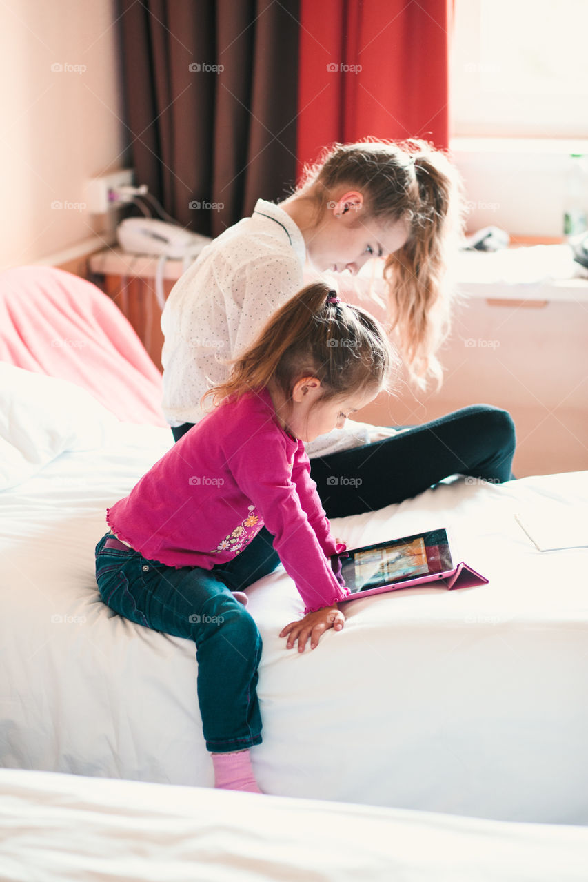 Teenage girl using mobile phone together with her little sister watching animated movie on tablet, both girls sitting in bed in bedroom