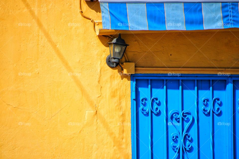 Yellow house wall with blue awning and blue door in sunlight 