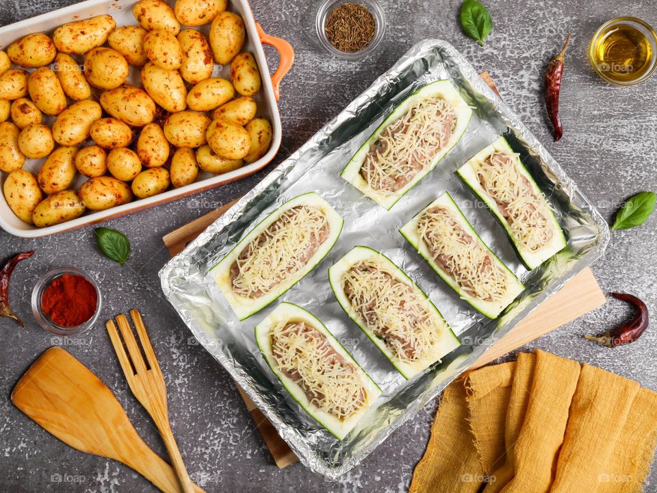 Beautiful view of green young zucchini stuffed with minced meat with grated cheese and small round potatoes in a dish with a napkin, spices and wooden cutlery lying on a dark stone background, flat lay close-up.