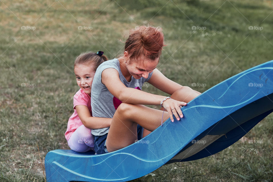 Teenage girl playing with her younger sister in a home playground in a backyard. Happy smiling sisters having fun on a slide together on summer day. Real people, authentic situations