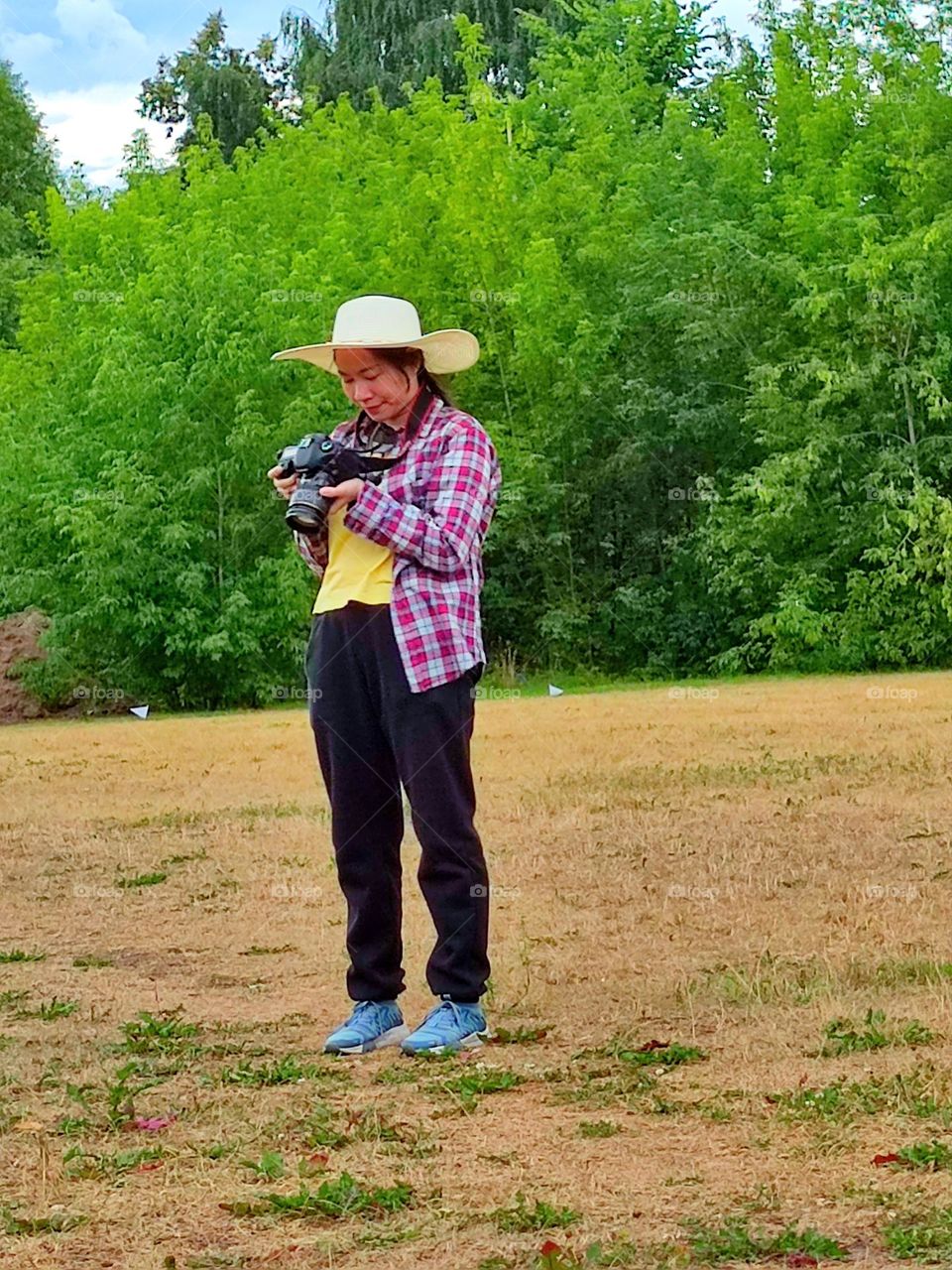 Summer day.  A field surrounded by green trees.  A girl in a straw hat stands on the field and looks into the photo camera