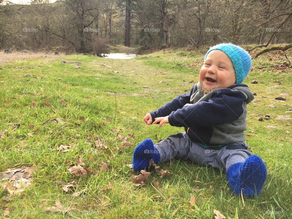 Happy baby boy wearing a blue stocking cap smiling for a photo sitting on grass out for a hike.