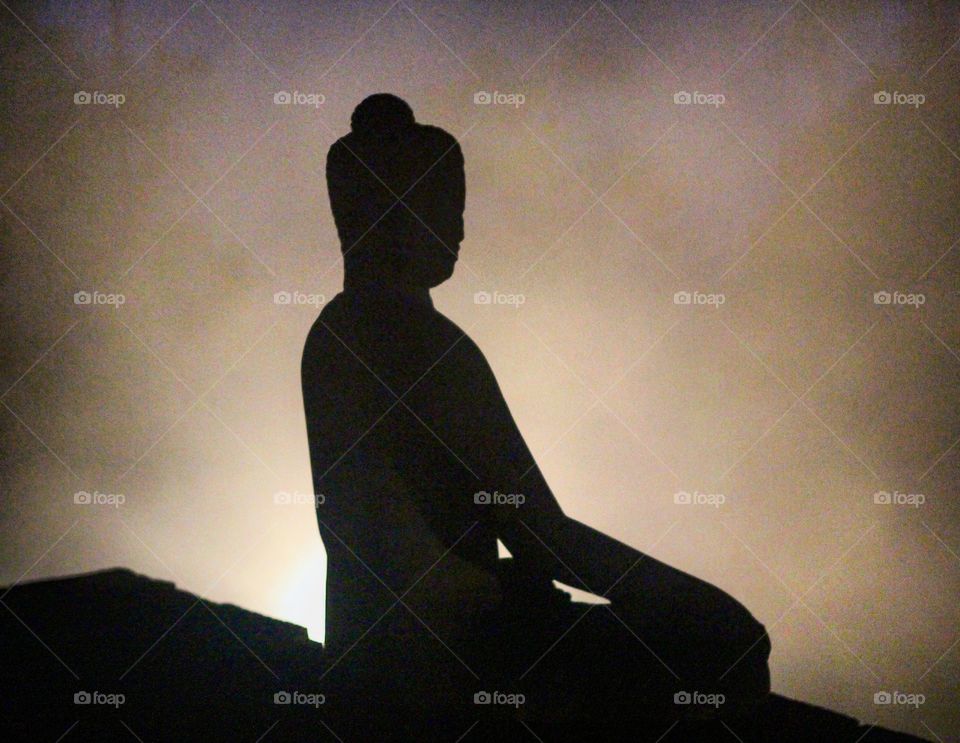 Silhouette of stone Buddha sitting in full lotus position, shrouded in mist and backlit with golden light. 