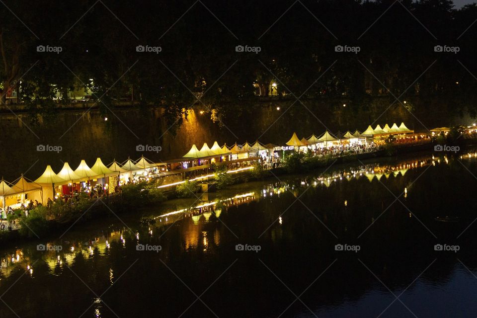 A portrair at night of the lights of an Italian market next to a river. the market lights and the stands themselves are reflected in the water and are lighting up the darkness.