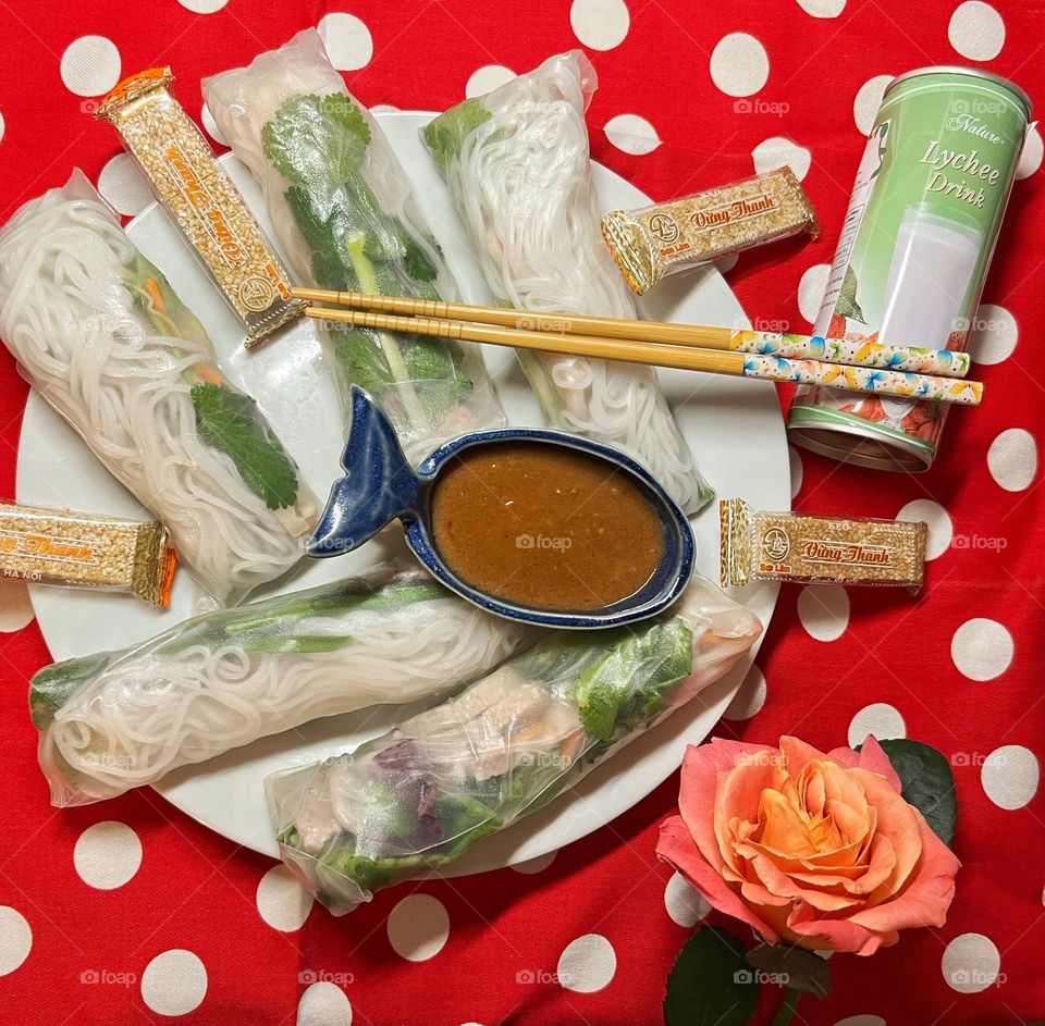 Vietnamese summer rolls on a polka dot underground with a dip, a lychee drink, sesame snack and a rose.