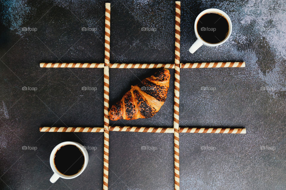 Businessman working morning with fresh delicious croissant and cup of hot black coffee on black background. Game of noughts and crosses. Top view, copy space, mockup. flat lay. creative concept.