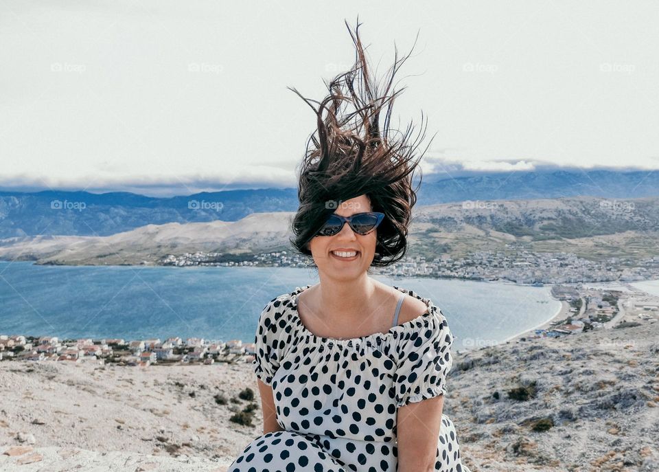 Smiling Caucasian woman in a polka dots flowy dress and sunglasses with hair blown up by the wind