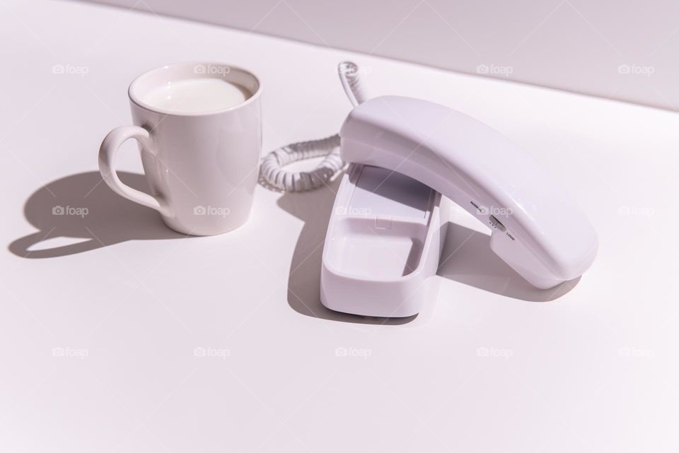 Hard light monochromatic white image of a white mug of milk and a white corded house telephone 