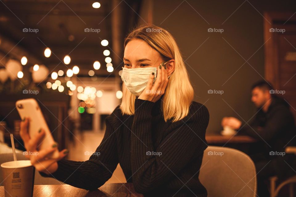 young woman sits in a mask.  blonde is sitting in a cafe and talking on the phone, quarantine time